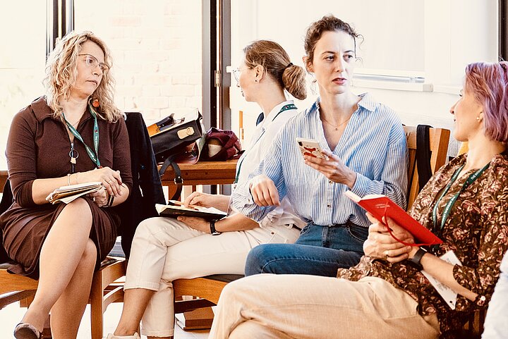 Four women are seated closely together in a brightly lit room, engaged in a discussion. One woman holds a phone and appears to be speaking, while the others listen intently, with two of them holding notebooks. The warm lighting and their focused interaction suggest a collaborative workshop setting.
